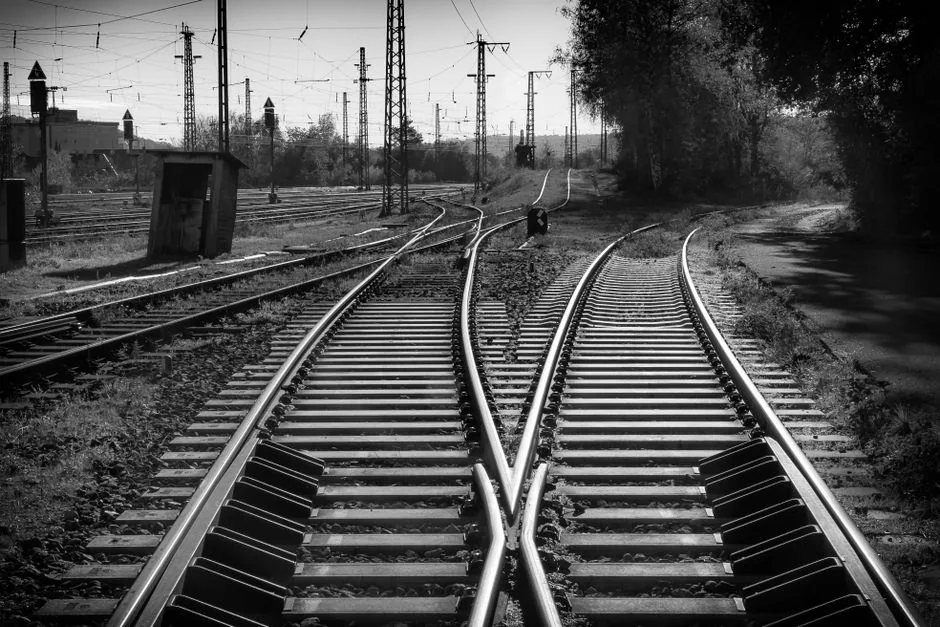 A black and white photograph of intersecting railway tracks with overhead wires and surrounding nature.
